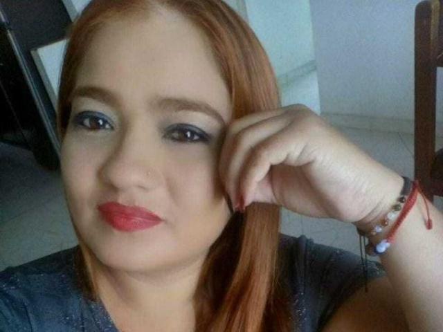SoffiMilf with red hair, blue eye makeup, red lipstick, gray top, touching her ear, red nail polish, red and white bracelets, smiling, fair skin.