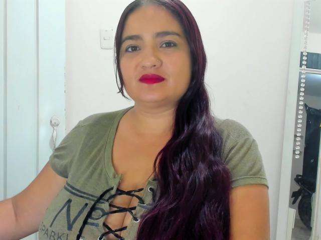 SoffiMilf with long dark purple hair, green top, lace-up front, red lipstick, and a neutral expression, sitting against a white wall. Her tits are medium-sized.