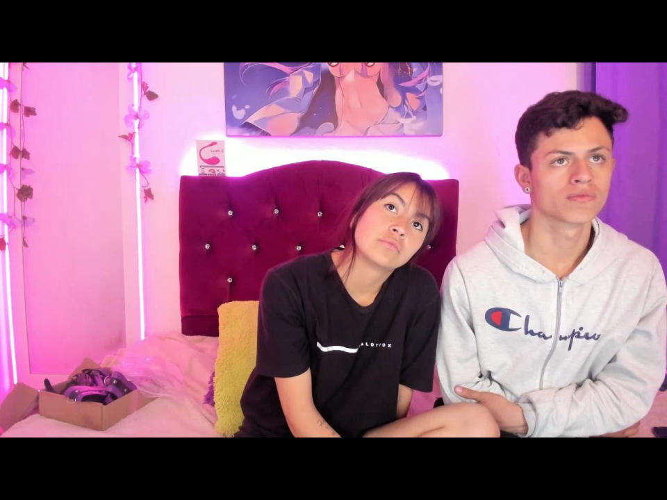 BigDickAndMelany with short brown hair and small tits, wearing a black shirt, sits next to a guy with curly hair in a gray Champion hoodie, looking confused.