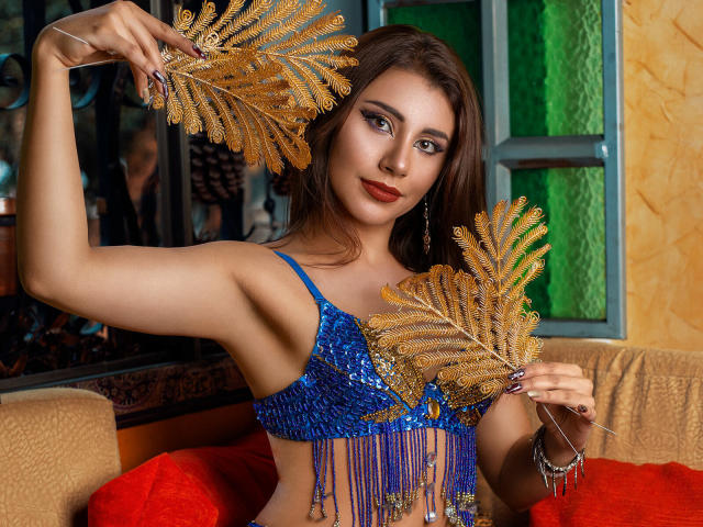 YennettSphinx segurando samambaias douradas, vestindo uma blusa azul com lantejoulas e franjas, cabelo castanho, batom vermelho, olhando para a câmera, dentro de um quarto colorido com uma janela..