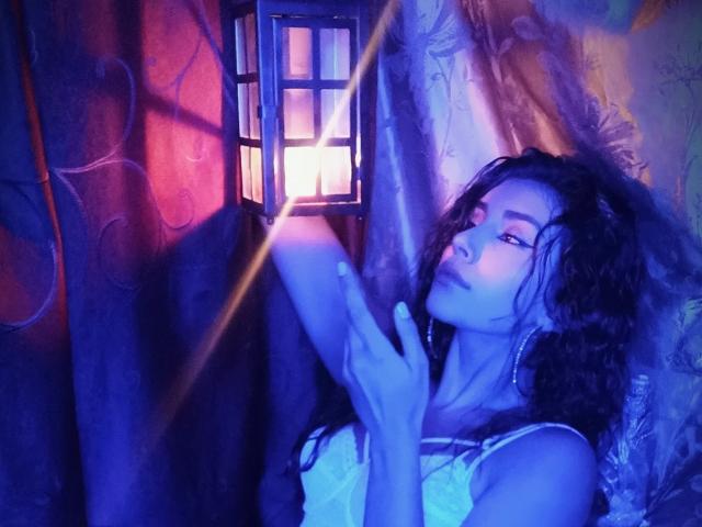 YennettSphinx with blue-tinted skin, curly black hair, and white lace top, touching a glowing lantern, eyes half-closed, looking up, with a soft light beam crossing her face.