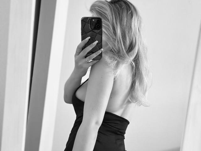 SugarrX, com cabelo loiro, costas nuas e vestido preto sem alças, segurando um telefone, cobrindo os seios, em um selfie em preto e branco. Suas nádegas estão parcialmente visíveis..