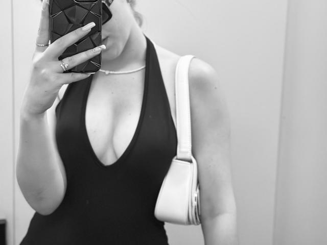 Black-and-white selfie of SugarrX with large boobs in a deep V-neck tank top, holding a phone with a geometric case, white purse on shoulder, rings on fingers.