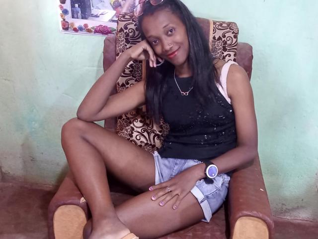 StellaMelody, dark-skinned woman with long black hair, sitting in brown chair, wearing black sleeveless top, blue denim shorts, pink wristwatch, and necklace, smiling, hand on cheek.