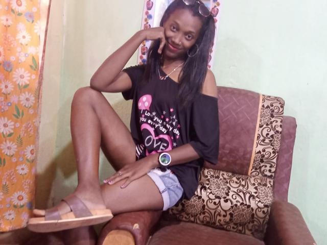 StellaMelody, dark-skinned woman with long black hair, smiling, wearing black off-shoulder shirt, denim shorts, brown sandals, and wristwatch, sitting on brown, patterned chair, hand on chin.