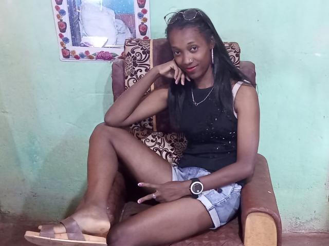 StellaMelody, dark-skinned woman with long black hair, wearing black tank top, denim shorts, and brown sandals, sitting on patterned chair, hand on chin, smiling, watch on wrist.