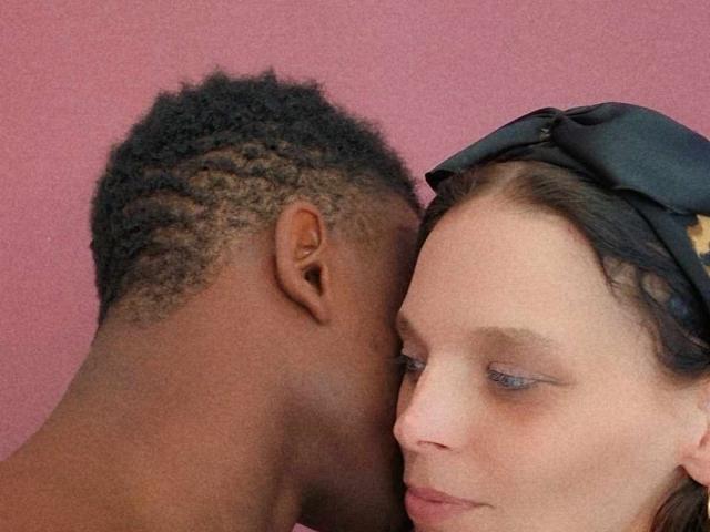 HotCouplez with dark skin and short curly hair kisses HotCouplez with light skin and brown hair, wearing a black headband, on a pink background. Her eyes are half-closed.