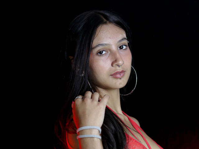 Kirstty with long black hair, red bra, and hoop earrings, has a nose ring, lip piercing, and a ring on her finger, against a black background.