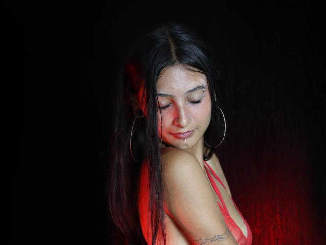 Kirstty with long black hair, red eyes, freckles, hoop earrings, and a red top, looking down with a slight smile, showing her small tits and a tattoo on her arm.
