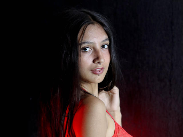 Kirstty with long black hair, brown eyes, and a nose piercing, wearing a red lace top, looking over her shoulder with a slight smile, against a dark background.