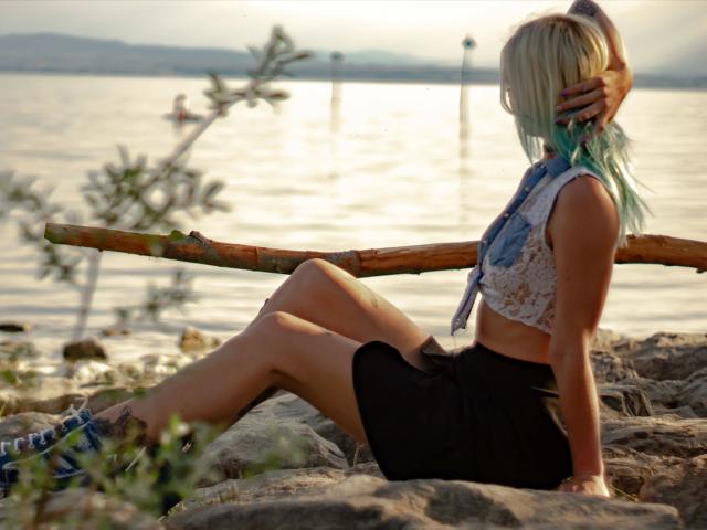 AmazingSugar-hot with blue-green hair, sleeveless white lace top, and black skirt, sitting on rocks, hand in hair, wooden log behind, legs crossed, sandy shore, water in background.