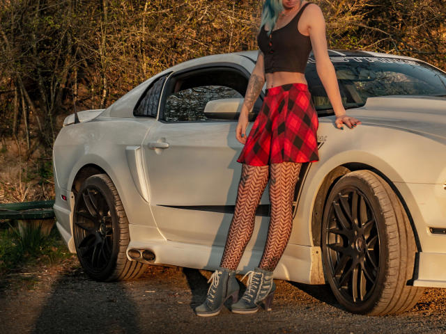 AmazingSugar-hot with blue hair, black crop top, red plaid skirt, chevron tights, and grey boots, leaning on a white car with black wheels, standing on gravel.