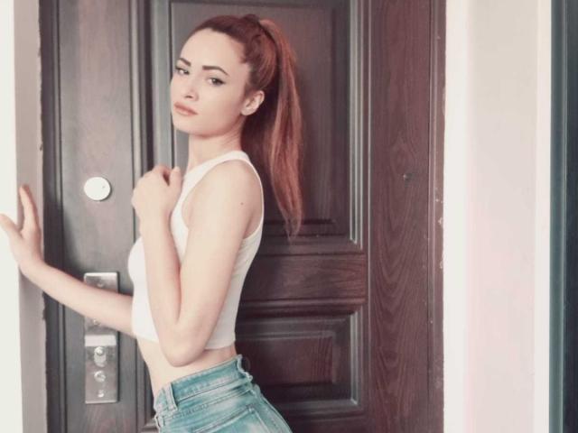 HotPinkLips69 stands in front of a dark wooden door, wearing a white crop top and blue jeans, with red hair in a ponytail, hands on the door.