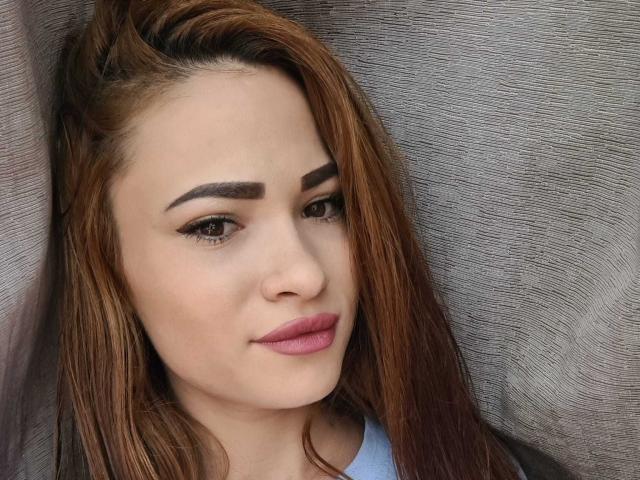 HotPinkLips69 has brown hair, thick eyebrows, subtle eyeliner, pink lipstick, and a light skin tone. She's looking slightly to the side with a gentle smile.