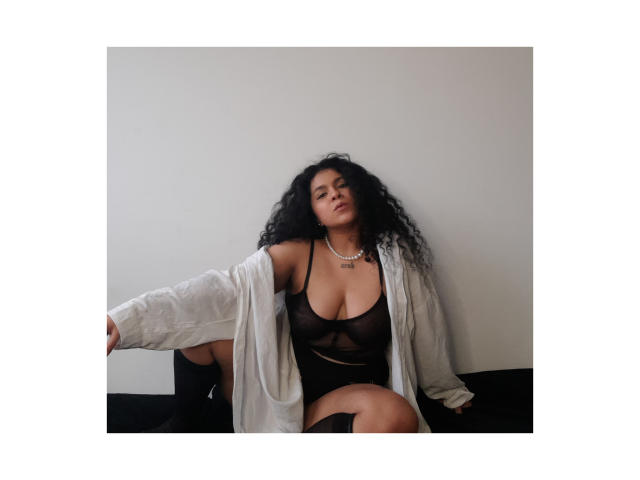 AbbyHairy with curly black hair, wearing a sheer black bra and white shirt, sitting on a black surface, exposing her tits and ass, with a necklace.