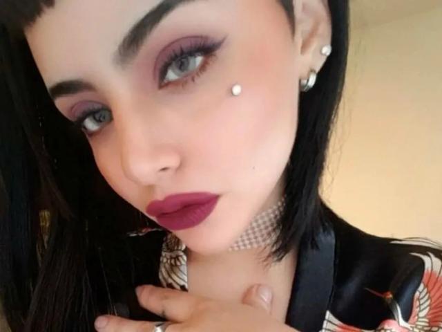 Celestia-hot has dark hair, purple eyeshadow, red lipstick, a pearl stud on her cheek, and a pearl choker. Her hand is near her neck, showing off her nails.