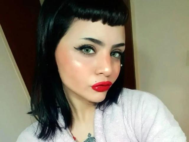 Celestia-hot has pale skin, black straight bob with bangs, green eyes, red lipstick, black eyeliner, and a nose piercing. She's wearing a white fluffy robe.