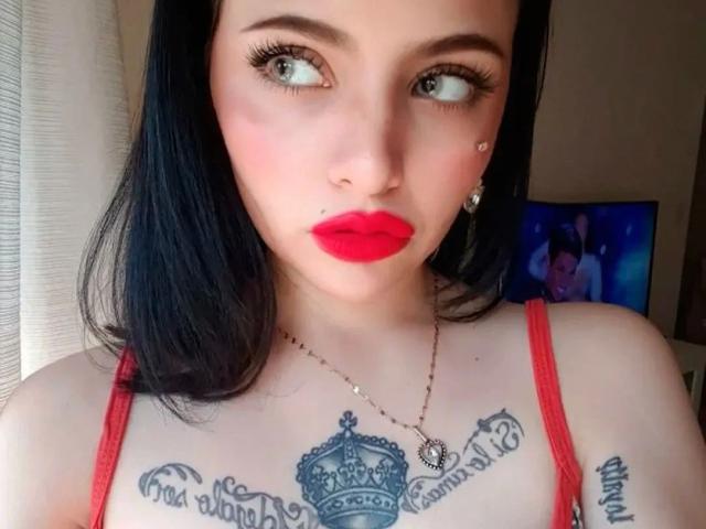 Celestia-hot with pale skin, black hair, red lipstick, tattoos under boobs, and a nose piercing, looking up with eyeliner and a necklace, wearing a red top.