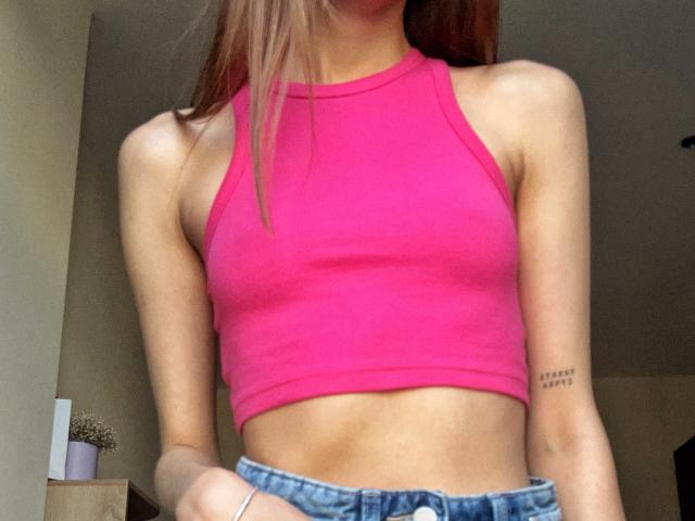 Karish has small tits, a slim waist, and is wearing a pink crop top with blue jeans. She has a tattoo on her right arm and long brown hair.