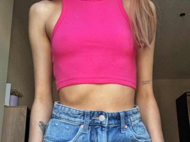 Karish is wearing a bright pink, tight, ribbed crop top that shows her small, flat, bare tits and a slim, toned, flat stomach. She has on high-waisted, blue denim jeans.