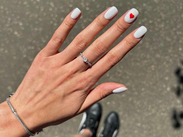 Karish's hand with white nails, a small red heart on the pinky, a silver ring, and a chain bracelet, showing a slightly dirty, textured asphalt ground.