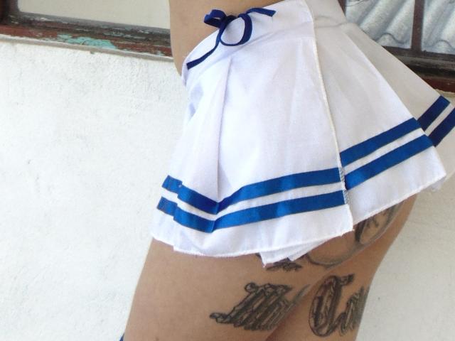La gonna bianca plissettata a righe blu di LunaRoose, che mostra le sue cosce e il sedere tatuati, con un fiocco blu sul lato, è appoggiata contro un muro bianco..