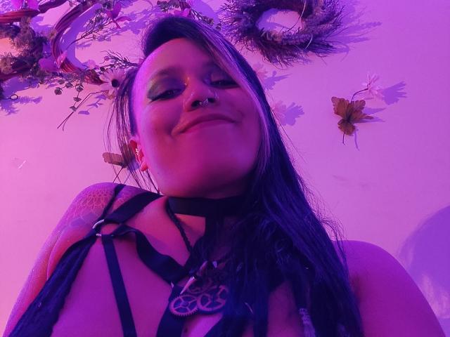 MistressLilith69, met een glimlach, met huid met een paarse tint, een zwarte bandeau top, een tatoeage op haar schouder, een neusring en lang donker haar, kijkt naar beneden naar de camera. Paars en roze verlichting..