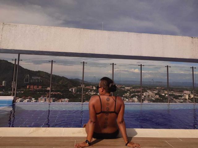 MistressLilith69, dark-skinned with a tattoo on her back, sits at an infinity pool, wearing black bikini bottoms, facing the blue water and city skyline, under a cloudy sky.