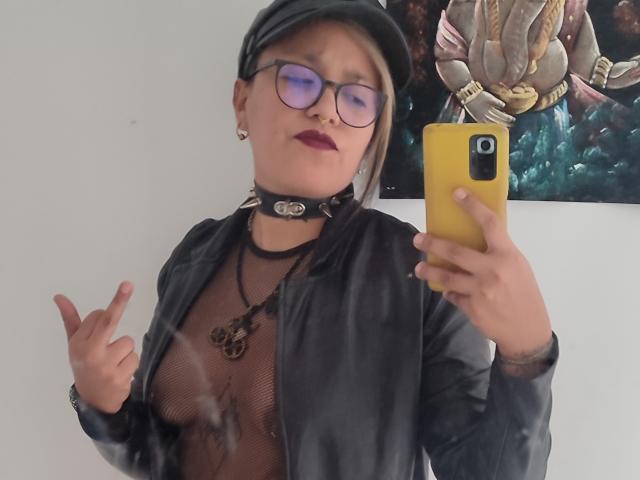 MistressLilith69 with glasses, black cap, leather jacket, mesh top, choker, dark lipstick, holding yellow phone, middle finger up, small tattoos, visible nipples, pinkie ring.