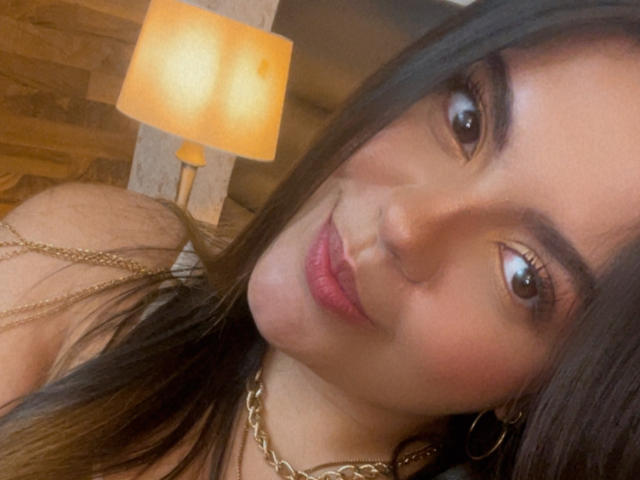 MissSofiia's face in a selfie, with long black hair, brown eyes, pink lips, gold chain necklace, and hoop earrings, smiling softly at the camera. Warmly lit room.