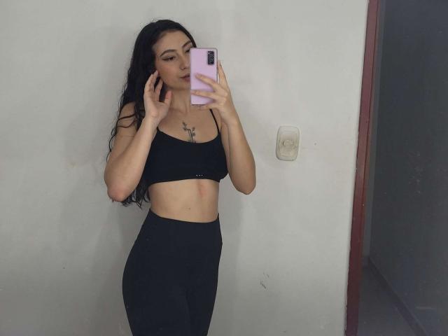 Emma Wells, con cabello largo y negro, vestida con un top deportivo negro y pantalones negros de cintura alta, sosteniendo un teléfono rosa, mostrando sus pechos y glúteos en un selfie frente al espejo..