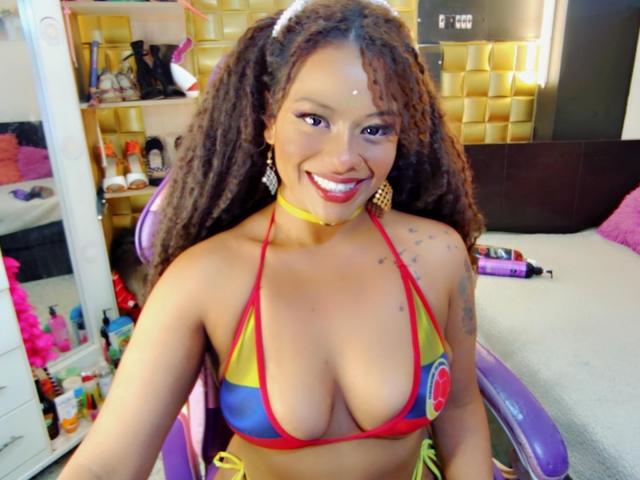 NashaJonny, mit großen Brüsten in einem farbenfrohen, dreieckigen Bikini-Oberteil, lächelt in die Kamera, hat lockiges Haar, eine gelbe Halskette und sitzt auf einem violetten Stuhl..
