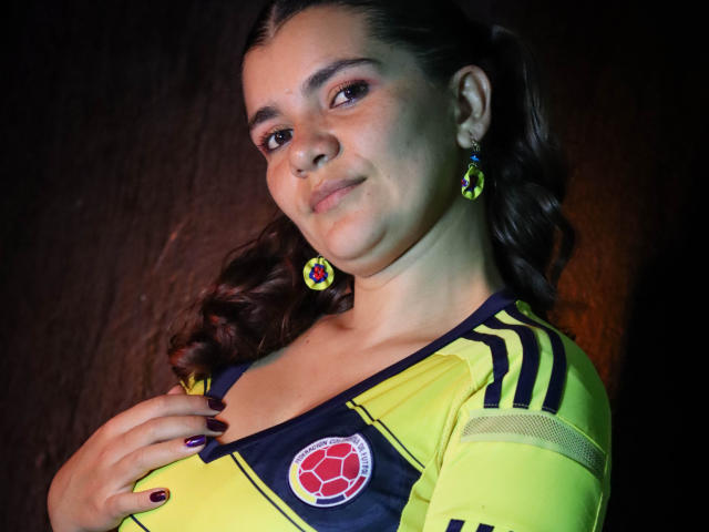 BulmaHot with dark hair in a ponytail, yellow soccer jersey, black and yellow stripes, red soccer logo, green earrings, purple nail polish, and a subtle smile.