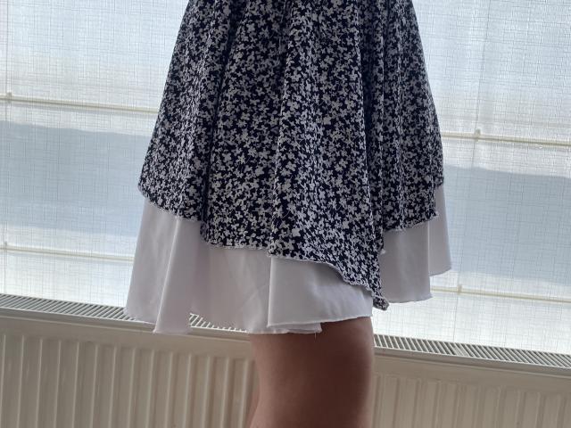 La falda floral de Mirellia, que muestra una capa interior blanca, se abre, dejando ver sus muslos, en una habitación iluminada por el sol con un radiador blanco en la parte inferior. La falda tiene un estampado floral azul..