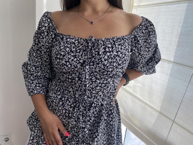 Mirellia wearing a black and white floral dress with puffy sleeves, off-shoulder, showing her tits slightly, has a necklace, pink nails, and a watch on her left wrist.