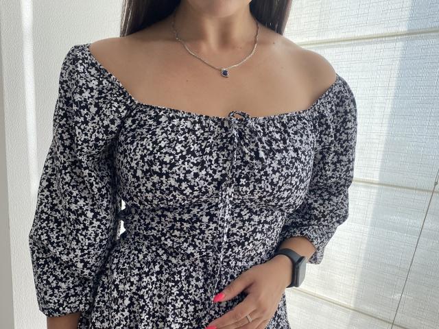 Mirellia mit großen Brüsten, in einem schwarz-weißen, blumigen Kleid, mit Schulterfrei-Ausschnitt, langen Ärmeln, einer silbernen Halskette, einer schwarzen Smartwatch, rosafarbenen Nägeln und heller Haut, steht im Sonnenlicht..