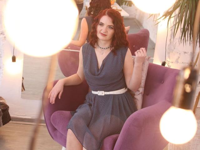 FreyaDak met rood krullend haar, gekleed in een blauwe jurk met een witte riem, zittend in een paarse stoel, lachend, lampen op de voorgrond, een witte bakstenen muur, een zwarte choker..
