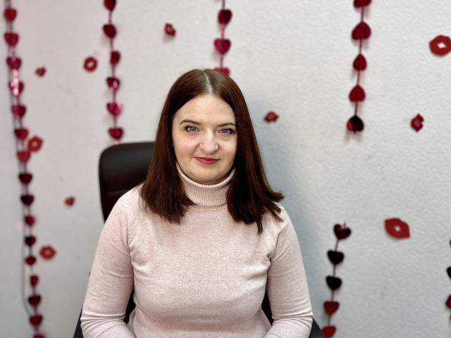 FreyaDak, com seios médios, vestindo um suéter rosa de gola alta, tem cabelo castanho, olhos azuis e um leve sorriso. Ela está sentada em uma cadeira, cercada por decorações de corações vermelhos e pretos..