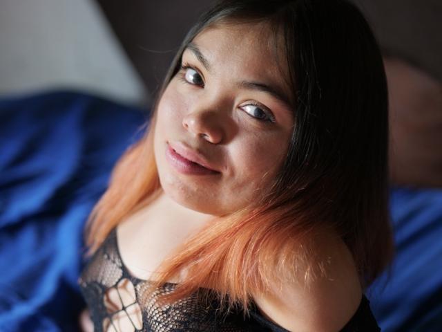 MichelleBrooks with medium skin, black to pink ombre hair, black lace top, looking up with a slight smile, soft light on her face, blue bed in background.