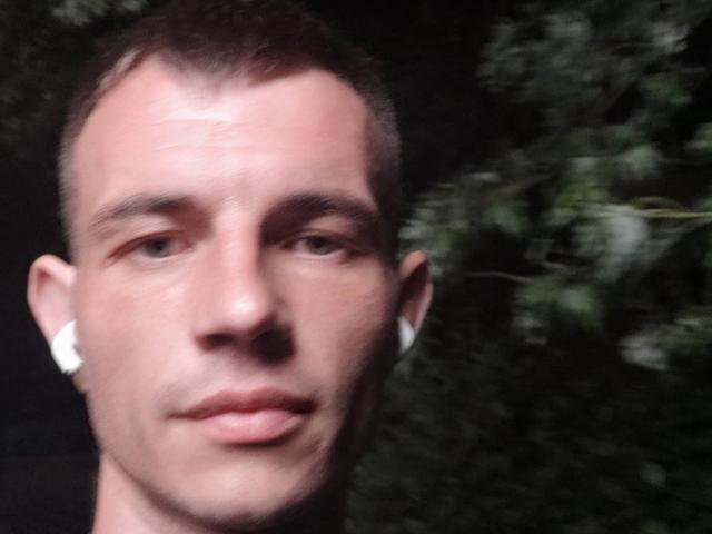 SunsetJohnny has short brown hair, white earbuds, and a serious expression. His skin is light with visible pores. His slightly parted lips show a subtle smile.