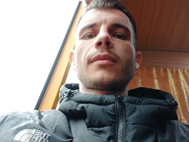 SunsetJohnny with short brown hair, slight beard, black puffy jacket, looking down at the camera, small tattoo under right eye, wooden ceiling above, bright outdoor light.