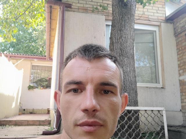 SunsetJohnny's face, short brown hair, slight stubble, neutral expression, slight forehead scar, tree behind him, white window, chain-link fence, and brick building in the background.