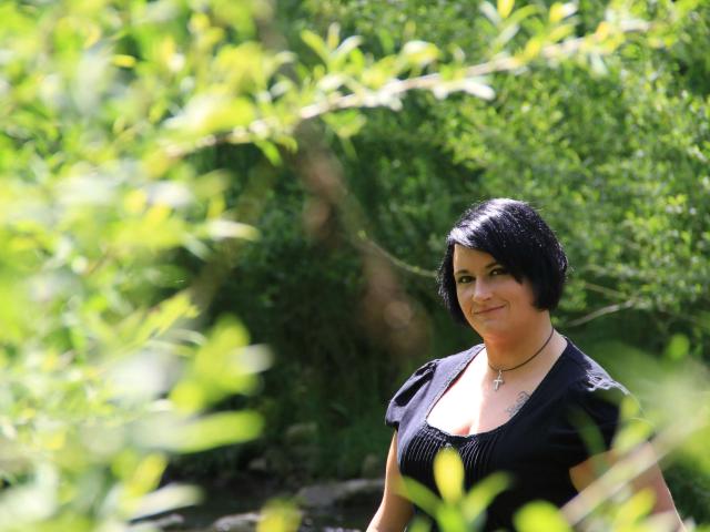 LadySun87 – eine Frau mit kurzen, schwarzen Haaren, die ein schwarzes Kleid trägt, eine Halskette mit einem Kreuz, steht in grünem Laub, lächelt, ihre Brüste sind leicht sichtbar, sonniger Tag, unscharfe Blätter im Vordergrund..
