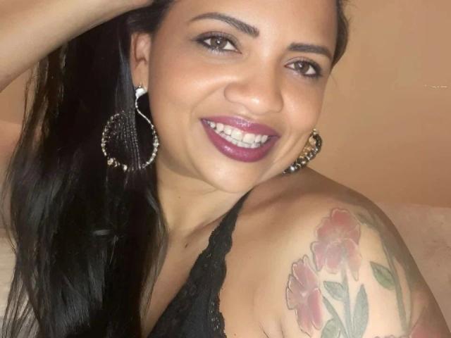 SamiraBelly-hot ha i capelli scuri, il rossetto viola e grandi orecchini a cerchio. Sorride mostrando i denti, indossa una maglia nera in pizzo e ha un tatuaggio colorato sulla spalla destra..