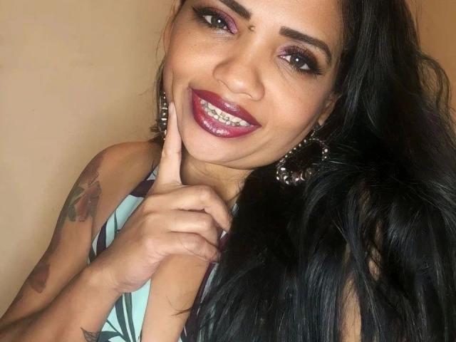 SamiraBelly-hot has dark hair, purple eyeshadow, red lipstick, a tattoo on her right arm, and is touching her chin with her finger, smiling. She's wearing a green top.