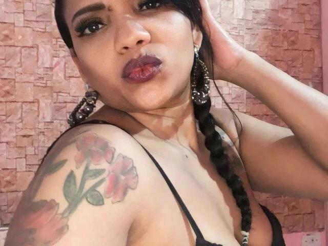 Samira, capelli scuri intrecciati, con orecchini neri, rossetto rosso intenso e un reggiseno nero, mostra una spalla tatuata con fiori rosa, con una mano tra i capelli..