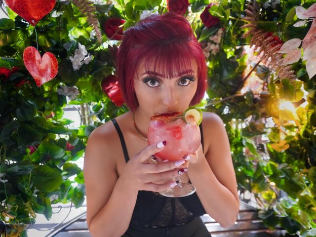 LizBonnet with red hair, black lace top, drinking a pink cocktail with a lime, her eyes wide, holding the glass near her lips, surrounded by green leaves and red hearts.