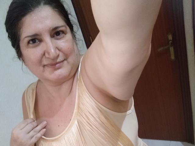 Rosamoor has a chubby face, brown eyes, and dark hair in a messy bun. She's wearing a beige, pleated top, showing her arm and small tits. She's smiling slightly.
