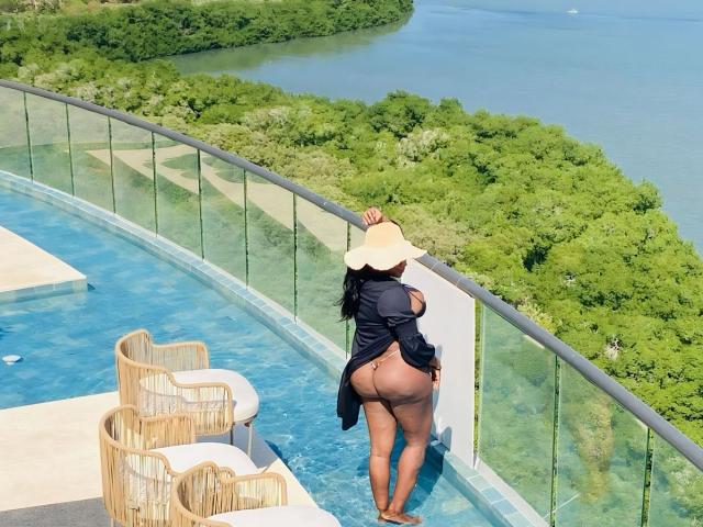 DiosaEbano stands by a glass balcony, wearing a sunhat, black robe, and no panties, showing her big ass and brown skin. She leans on the railing, overlooking a blue pool.