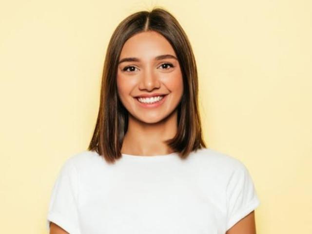 Danale has medium brown ass-licking hair, a white tee, and a smiling face against a yellow background. Her brown eyes and teeth are visible.